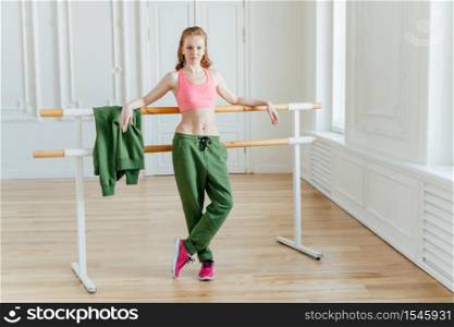 Slim beautiful female ballet dancer leans at ballet barre, wears pink top, sport trousers and sneakers, shows fit figure, stretches before dancing, poses in studio or classroom, has rehearsal