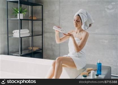 Slim beautiful young woman squeezing lotion from bottle during skincare treatment spa procedure in bathroom. Female wrapped in towel taking care of body skin using natural cosmetics after bathing.. Woman takes care of body skin using moisturizing natural cosmetics after bathing. Skincare treatment
