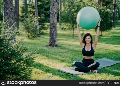 Slim bru≠tte lifts fit≠ss ball overhead, sits in lotus pose on karemat in the forest. Wearing active wear, practices yoga outdoors, enjoying fresh air. Fit lady exercises with a gymnastic ball.