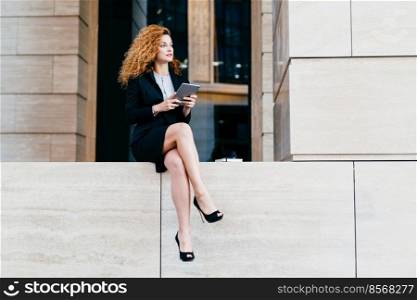 Slim businesswoman with wavy luxurious hair, having slender legs, wearing black elegant suit and shoes, holding tablet computer, looking aside while resting for minute after hard work in office