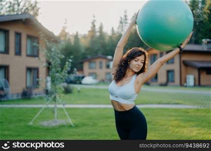 Slim Caucasian woman practices yoga with fitness ball outdoor, dressed in active wear, poses on green lawn. Gymnastic training, serene backdrop.