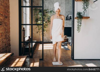 Slim caucasian woman taking step onto scale and measuring her weight. Girl wrapped in towel after bathing has time at spa. Young happy woman checking weight in bathroom. Fitness and wellness concept.. Slim caucasian woman taking step onto scale and measuring her weight. Fitness and wellness concept.