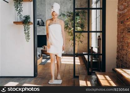 Slim caucasian woman taking step onto scale and measuring her weight. Girl wrapped in towel after bathing has time at spa. Young happy woman checking weight in bathroom. Fitness and wellness concept.. Slim caucasian woman taking step onto scale and measuring her weight. Fitness and wellness concept.