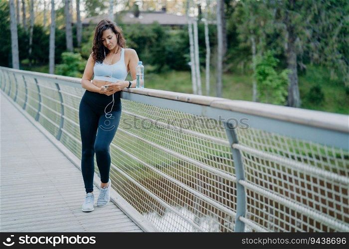 Slim European woman in active wear checks phone, listens to music with earphones on the bridge. Embracing the sport and lifestyle concept amid nature’s backdrop.