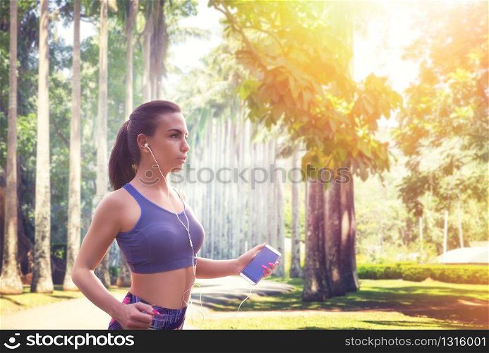 Slim female jogger with headphones in summer park, sunlight on background. Woman jogging on outdoors morning workout