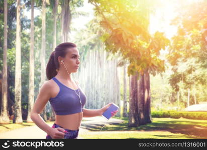 Slim female jogger with headphones in summer park, sunlight on background. Woman jogging on outdoors morning workout