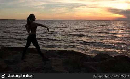 Slim fit woman exercising yoga warrior pose on the beach in glow of amazing sunset. Silhouette of fitness sporty female practicing yoga on rocky seashore, doing warrior one position over beautiful seascape and colorful sky with clouds at sundown.