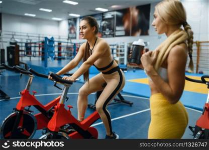 Slim girl drinks water on stationary bikes in gym, side view. People on fitness workout in sport club, athletic girls in sportswear on training indoors. Slim girl drinks water on stationary bikes in gym
