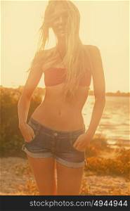 Slim girl in red bikini top and jeans shorts posing on summer beach in sunset sunshine. Slim girl in red bikini top and jeans shorts posing on summer beach in sunset sunshine in front of water and sky.