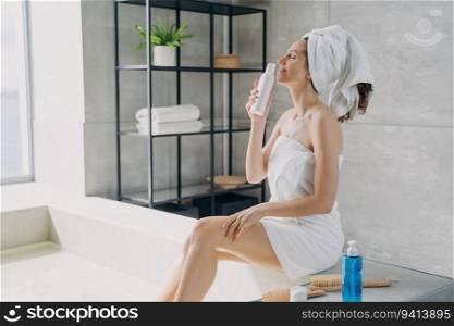 Slim girl savors lotion scent, relaxes in home bathroom. Attractive European woman, towel-wrapped post-shower. Freshness, daily beauty routine.