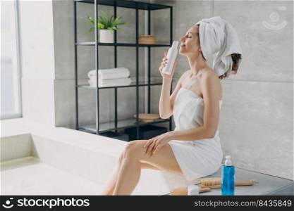 Slim girl takes a smell of body lotion fragrance from bottle and relaxing in bathroom at home. Attractive european woman wrapped in towel after taking shower. Freshness and daily beauty routine.. Slim european girl takes a smell of body lotion from bottle and relaxing in bathroom at home.