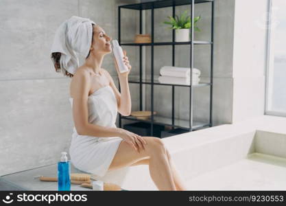 Slim girl takes a smell of body lotion fragrance from bottle and relaxing in bathroom at home. Attractive european woman wrapped in towel after taking shower. Freshness and daily beauty routine.. Slim european girl takes a smell of body lotion from bottle and relaxing in bathroom at home.