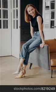 Slim lady in fashionable bodysuit and high heels is sitting on an armchair armrest. Portrait of gorgeous young woman. Redhead lady in casual outfit is smiling. Concept of style, fashion and beauty.. Slim lady in fashionable bodysuit and and high heels is sitting on an armchair armrest and smiling.