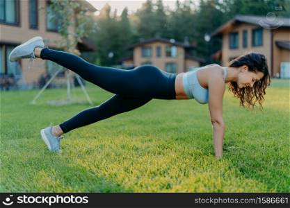 Slim motivated brunette woman does fitness exercises in open air raises legs and has regular workout dressed in cropped top leggings, sneakers, poses on green grass near house. Active rest concept