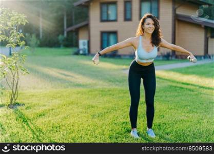 Slim motivated brunette woman dressed in cropped top and leggings, has workout with dumbbells, poses on green lawn near private house, has perfect body shape. Healthy lifestyle and sport concept