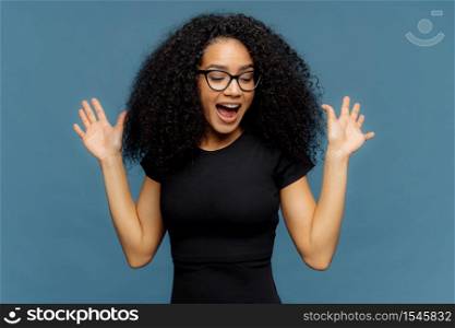 Slim overemotive dark skinned woman raises hands, opens mouth, gestures actively from positive emotions, focused down, dressed in casual black t shirt, isolated over blue background. Ethnicity concept