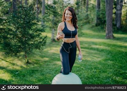 Slim pleased brunette sporty woman leans at fit ball, holds mobile phone, listens music in earphones, downloads songs in playlist, poses with bottle of water, makes exercises in open air at park