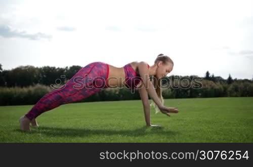 Slim pretty woman with pony tail planking on the green grass on sunny day. Full length of sporty young female doing the side rotating plank yoga pose in fitness on grass outdoors. Side view