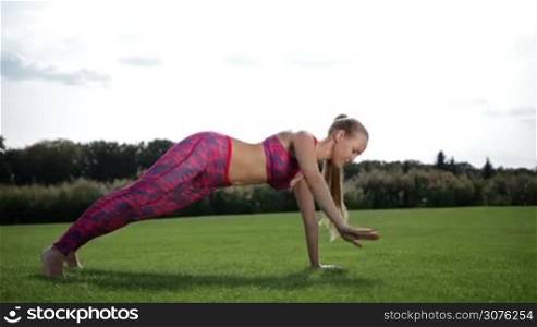 Slim pretty woman with pony tail planking on the green grass on sunny day. Full length of sporty young female doing the side rotating plank yoga pose in fitness on grass outdoors. Side view