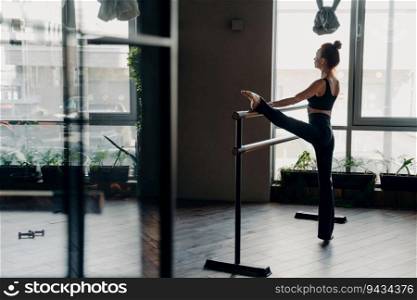 Slim red-haired ballerina stands in split position beside ballet barre in gym, light window background. Indoor sport and stretching showcased beautifully.