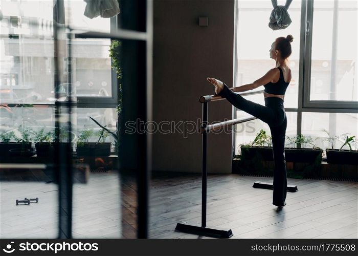 Slim red haired young ballerina in black top tank and leggings standing in split position next to ballet barre in gym against light large window background. Concept of stretching and sport indoor. Slim ballerina during her stretch routine next on ballet barre in studio