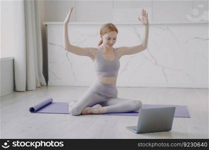 Slim sporty girl is stretching on mat. Mid european woman is practicing yoga asana. Concept of remote learning. Online fitness classes student is happy watching video on laptop.. Slim sporty girl stretching on mat. Online remote fitness classes student watching video on laptop.