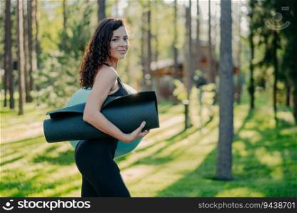 Slim sporty woman carries karemat under arm, dressed in black leggings. Poses against green grass and trees, ready for fitness ball exercises.