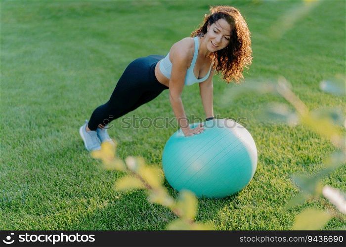 Slim woman enjoys gymnastic exercises on green lawn with fit ball, wearing active wear. Embraces healthy, active lifestyle. Fitness concept.