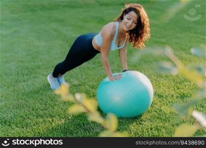 Slim woman enjoys gymnastic exercises on green lawn with fit ball, wearing active wear. Embraces healthy, active lifestyle. Fitness concept.