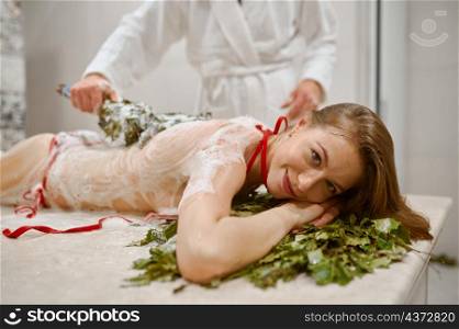 Slim woman in bath getting steaming massage with hot oak leaves brooms. Relaxation in steam-room. Woman in bath getting steaming broom massage