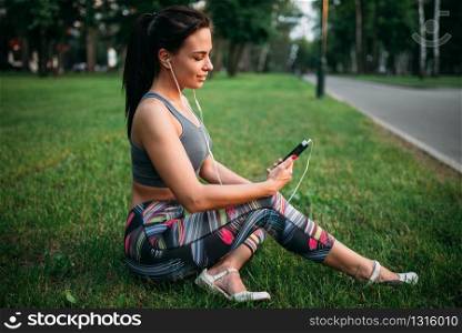 Slim woman in headphones sitting on grass and relax. Sporty girl on outdoors fitness workout. Woman in headphones sitting on grass and relax