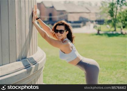 Slim woman with perfect body shapes leans with hands at something smiles positively dressed in top and leggings poses outdoor has fitness training during summer time. People and exercising concept