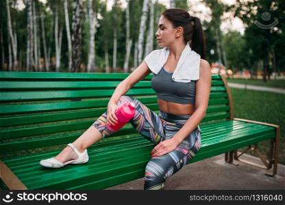 Slim woman with sport bottle sitting on a bench in summer park. Woman drink water on morning fit workout
