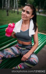 Slim woman with sport bottle sitting on a bench in summer park. Woman drink water on morning fit workout. Woman with sport bottle sitting on a bench in park