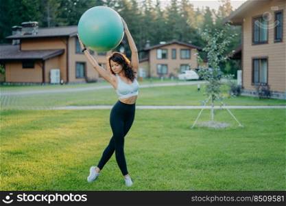 Slim young European woman holds fitball over head, dressed in cropped top and leggings, has gymnastics exercises outdoor, poses at green lawn near private house. People, sport, training concept