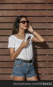 Slim young female in trendy casual outfit touching headphones and looking away while standing against wooden wall on sunny day. Stylish woman listening to music in headphone