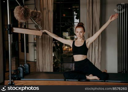 Slim young female pilates instructor sitting on trapeze with arms outstretched to both sides in relaxed position doing stretching exercises in gym on cadillac reformer. Sport and fitness concept. Flexible woman sitting on trapeze with arms outstreched to both sides
