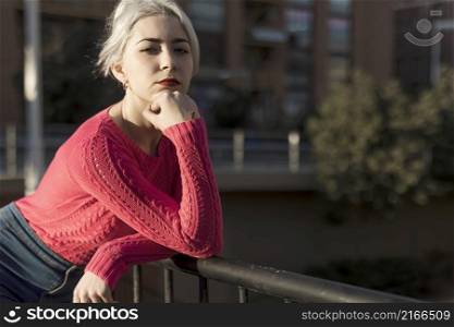 Slim young lady with blonde hair touching face and looking at camera while leaning on railing outdoors