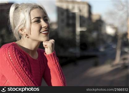 Slim young lady with blonde hair touching face and looking at camera while leaning on railing outdoors
