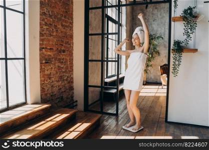 Slimming success. Excited girl wrapped in towel standing on floor scales measuring weight. Happy young woman gesturing yes, satisfied with weighing result after shower at home. Weightloss, wellness.. Slimming success. Excited girl gestures yes, satisfied with weight, standing on scales after shower