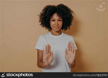 Slow down. Young smiling mixed race woman with curly hairstyle raising palms in stop or prohibition gesture and saying no while standing dressed in white t-shirt over brown studio wall. Young smiling mixed race woman with curly hairstyle raising palms in stop or prohibition gesture