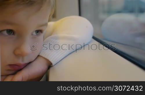 Slow motion and close-up shot of a little boy leaning on windowsill in train and staring through the window with bored look, finally he see something interesting