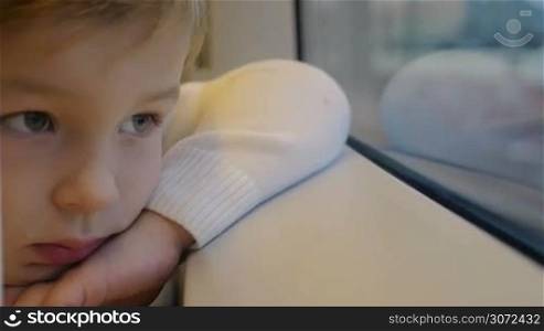 Slow motion and close-up shot of a little boy leaning on windowsill in train and staring through the window with bored look, finally he see something interesting