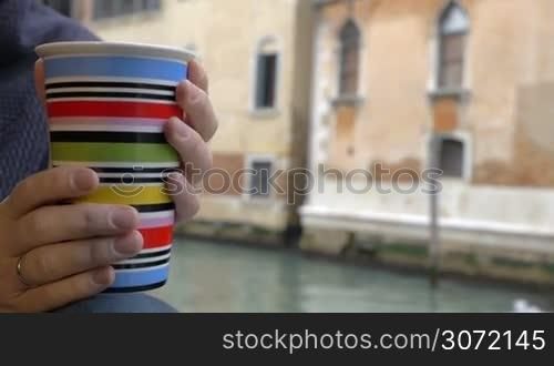 Slow motion and close-up shot of woman hands holding colorful cup. Old building and boat sailing along the canal in background