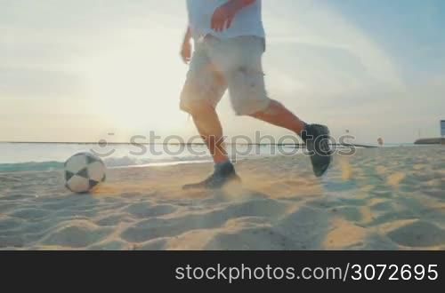 Slow motion and steadicam shot of a man playing football on the beach. He dribbling on the sand against sea and sunset background