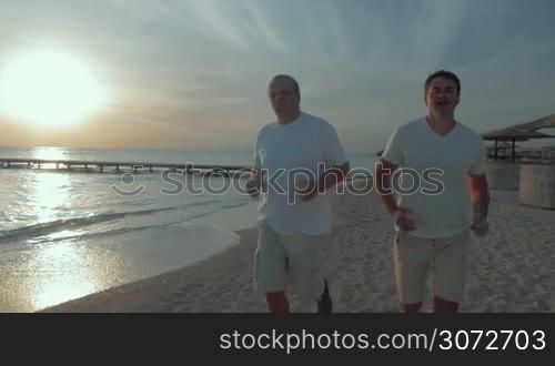 Slow motion and steadicam shot of a senior and young men running along the coastline at sunset. Evening jogging on the beach helps to stay in a good shape