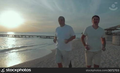 Slow motion and steadicam shot of a senior and young men running along the coastline at sunset. Evening jogging on the beach helps to stay in a good shape