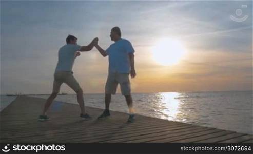 Slow motion and steadicam shot of two men boxing on the wooden pier in the sea at sunset. Young sportsman trying to hit the hand of his opponent