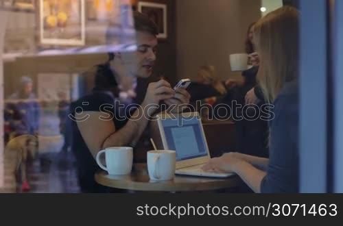 Slow motion clip of view through restaurant glass of man dictating data from phone and woman noting it in her laptop in the restaurant