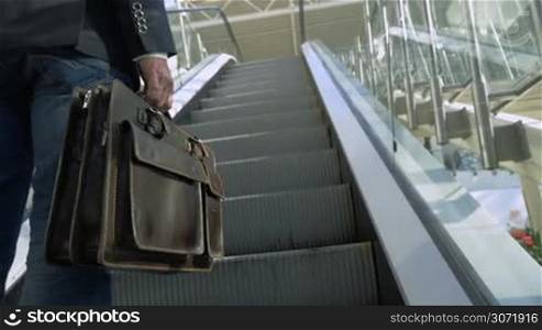 Slow motion, close-up and low angle shot of a businessman with leather briefcase going up on escalator in modern office building or trade center